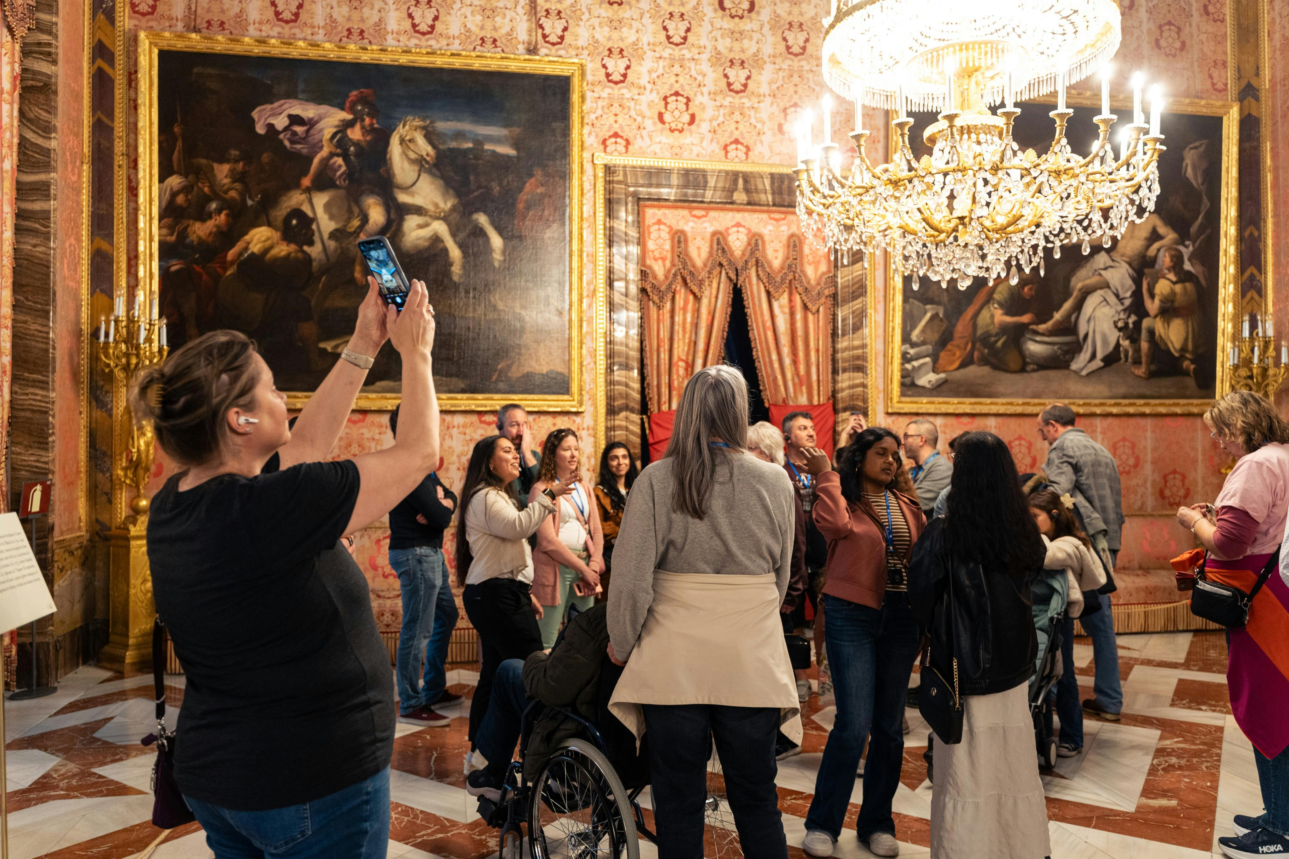 A group of people in a lavishly decorated room with gold-framed paintings, ornate wallpaper, and a large chandelier.