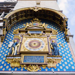 En utsmyckad astronomisk klocka med guld- och blådetaljer, statyer och romerska siffror, monterad på en arkitektonisk struktur.
