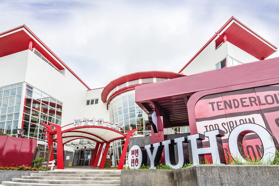 A modern building with red and white accents, featuring a large glass facade and a prominent 'YUH HO' sign in front.