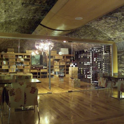 Vinkällare i sten med trägolv, bord, mönstrade stolar och vinställ. En utsmyckad ljuskrona hänger från det välvda taket.