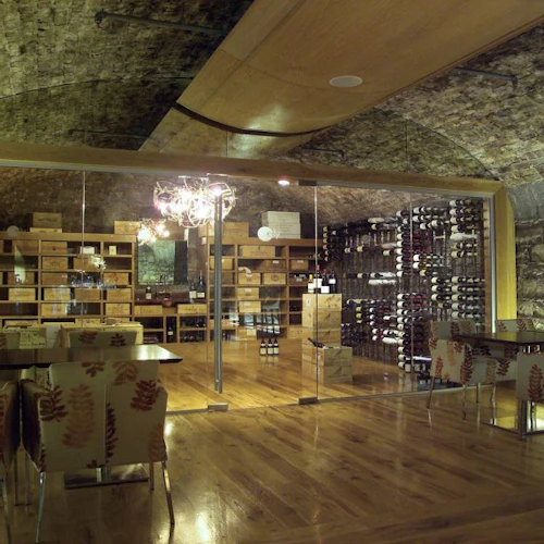 Stone wine cellar with wooden floors, tables, patterned chairs, and wine racks. An ornate chandelier hangs from the arched ceiling.