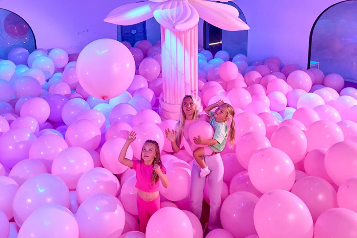Una dona sosté un nen envoltat de globus roses, amb un altre nen al seu costat, en una habitació il·luminada de color rosa amb una gran columna.