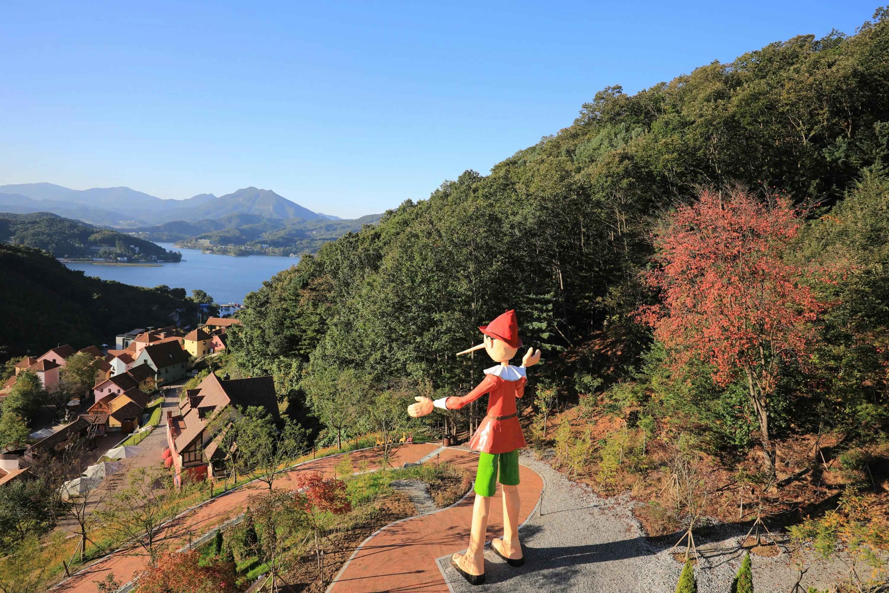 A statue of Pinocchio overlooking a scenic landscape with trees, a path, and a lake in the distance under a clear sky.
