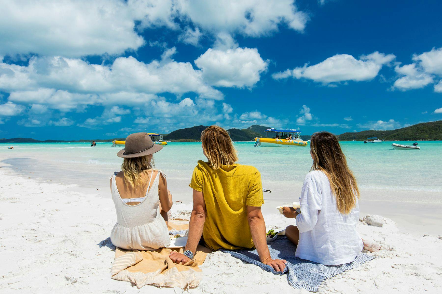 Three people sitting on a sandy beach, facing the ocean with boats and mountains visible under a bright blue sky.