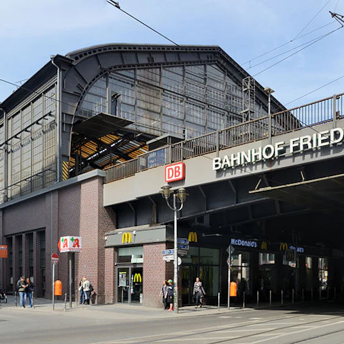 Upptagen stadsgata med människor som går och cyklar framför Bahnhof Friedrichstrasse tågstation. Spårvagnar och butiker är synliga.