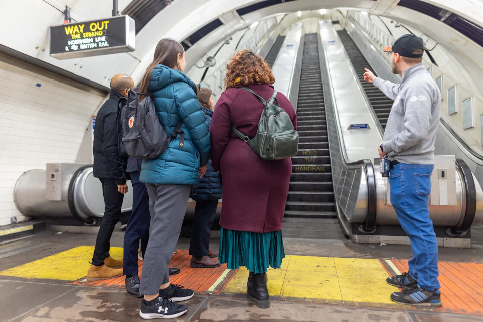 A group of people standing at the base of an escalator in a subway or train station with signage visible above.