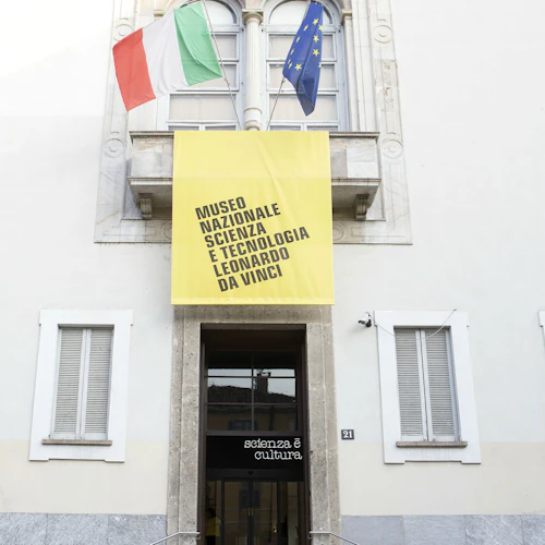 Fasad av en byggnad med italienska och EU-flaggor, en gul banderoll med texten "Museo Nazionale Scienza e Tecnologia Leonardo da Vinci."