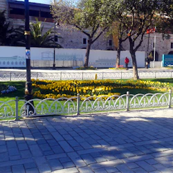 En park med gula blommor, dekorativa metallstaket och träd bredvid en stenbyggnad under en klar himmel. Folk går i närheten.