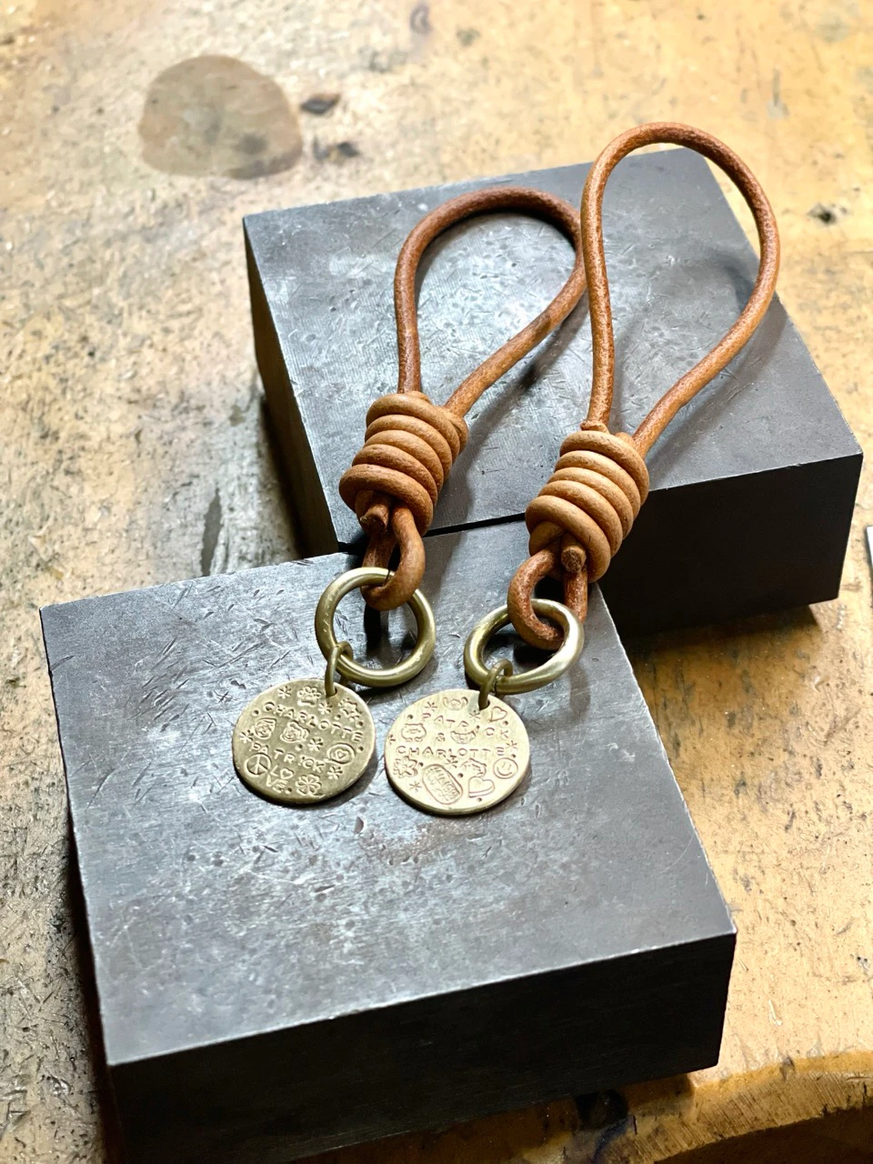 Two brown leather keychains with circular metallic charms placed on dark metal blocks on a wooden surface.