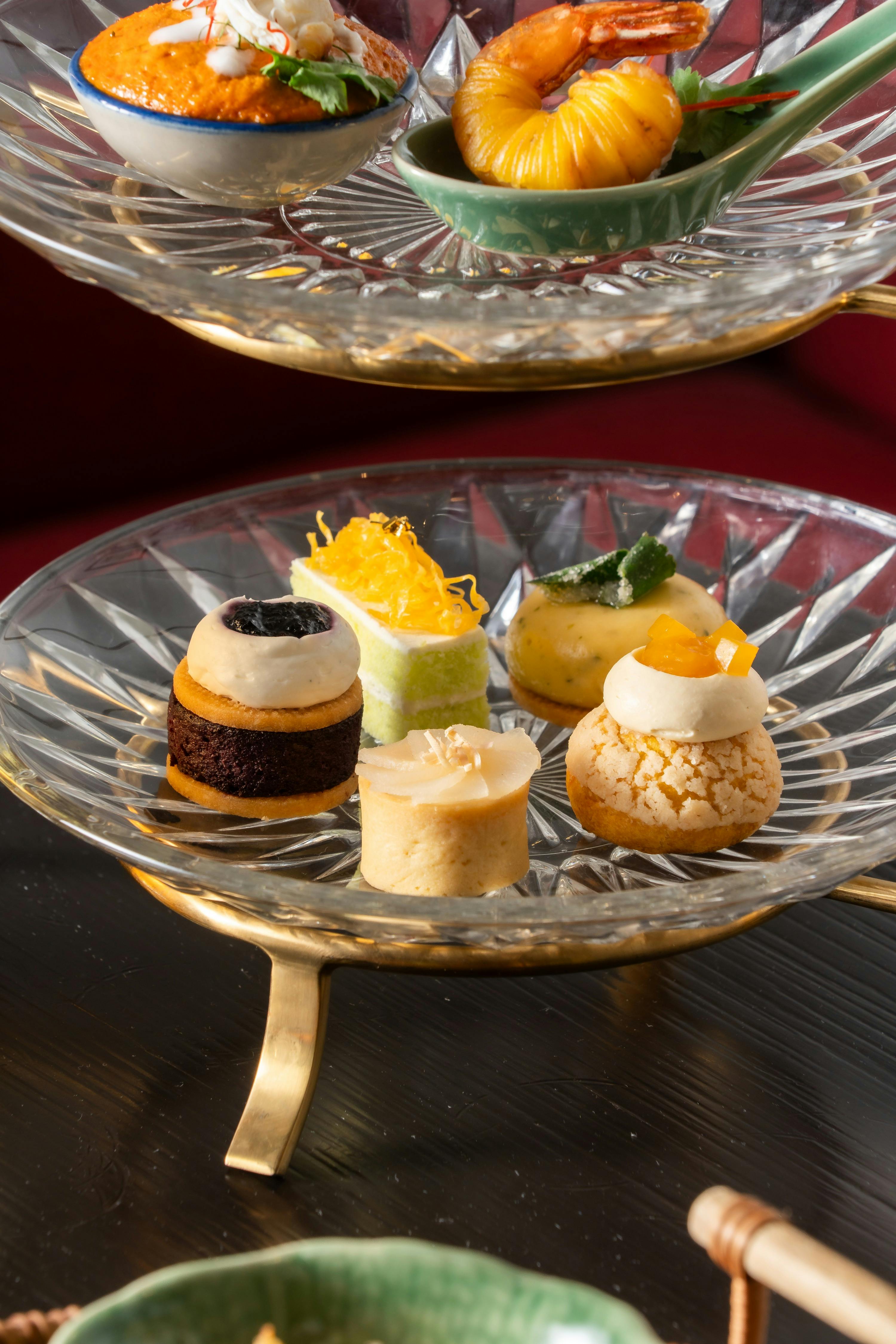 A glass tray on a golden stand holds five assorted bite-sized desserts with colorful toppings, placed on a dark surface.