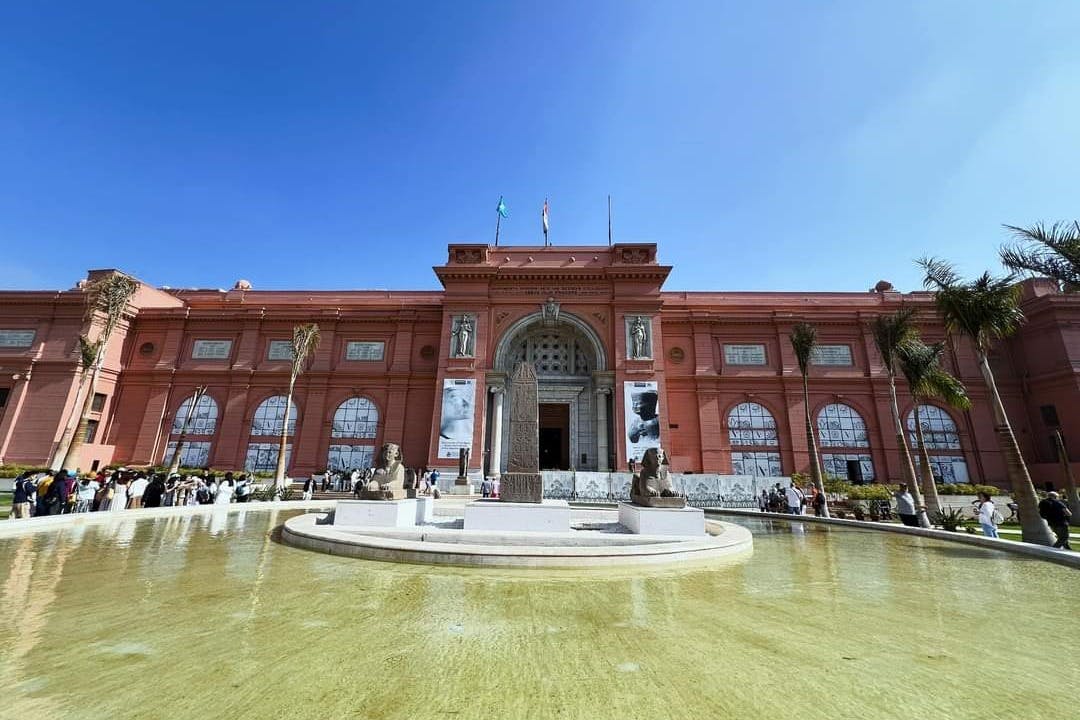 红色建筑，设有拱形入口、柱子和雕像，围绕着长方形泳池前的中央方尖碑。 上方晴朗的蓝天。