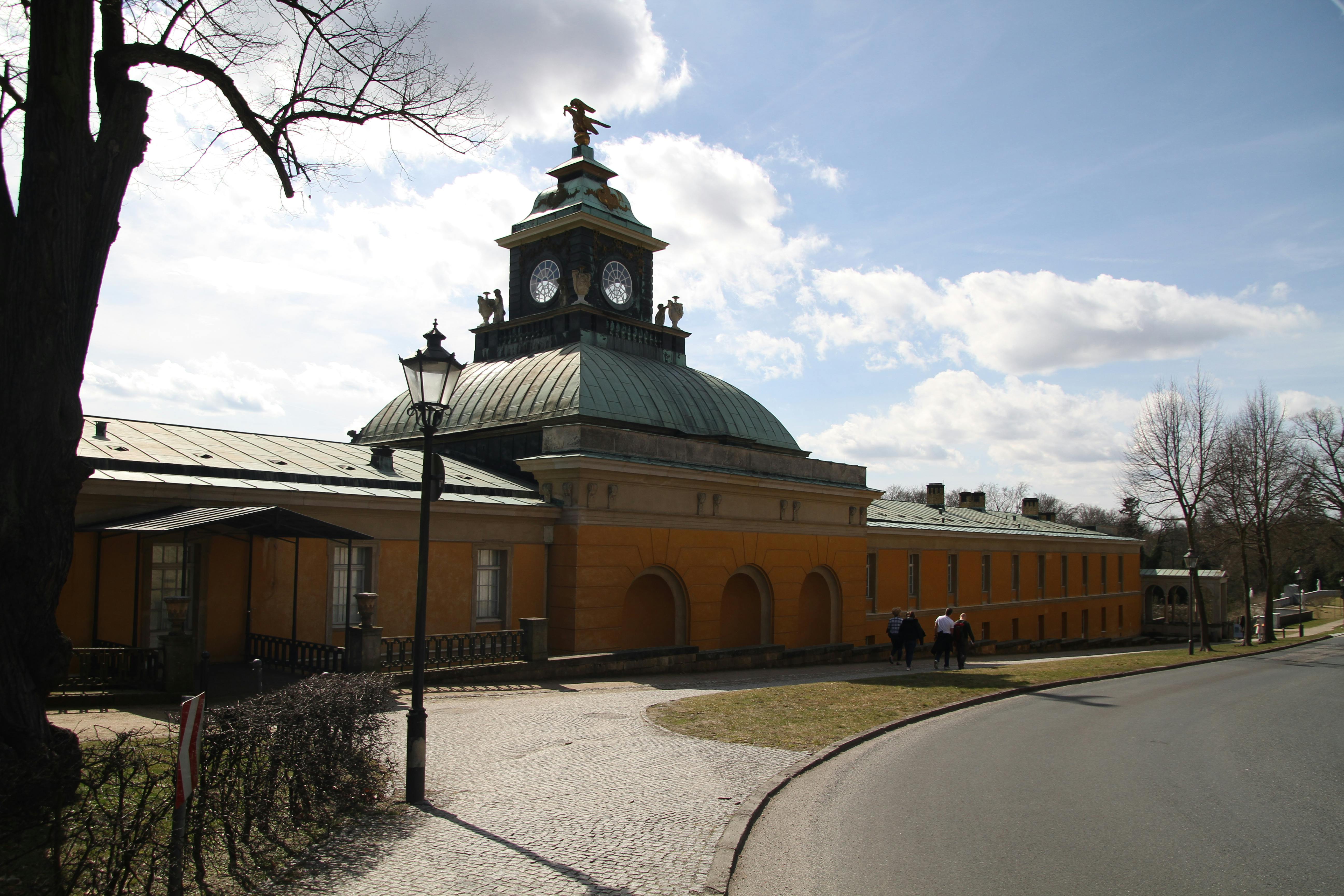 Un edifici de color groc mostassa amb torre de rellotge i cúpula verda, vorejat per un camí empedrat i arbres, sota un cel parcialment ennuvolat.