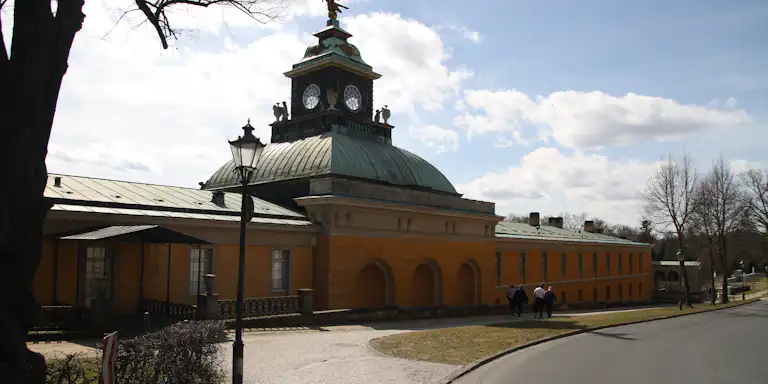 Ein senfgelbes Gebäude mit einem Uhrenturm und einer grünen Kuppel, umrahmt von einem Kopfsteinpflasterweg und Bäumen, unter einem teilweise bewölkten Himmel.
