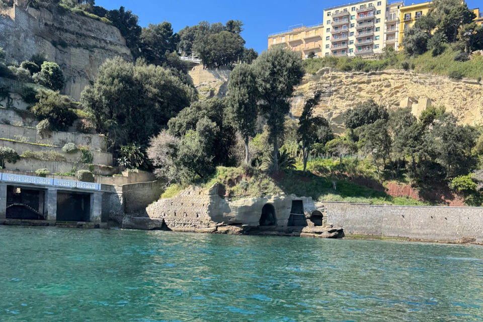 A rocky shoreline with caves, trees on a hillside, and a residential building in the background under a clear blue sky.