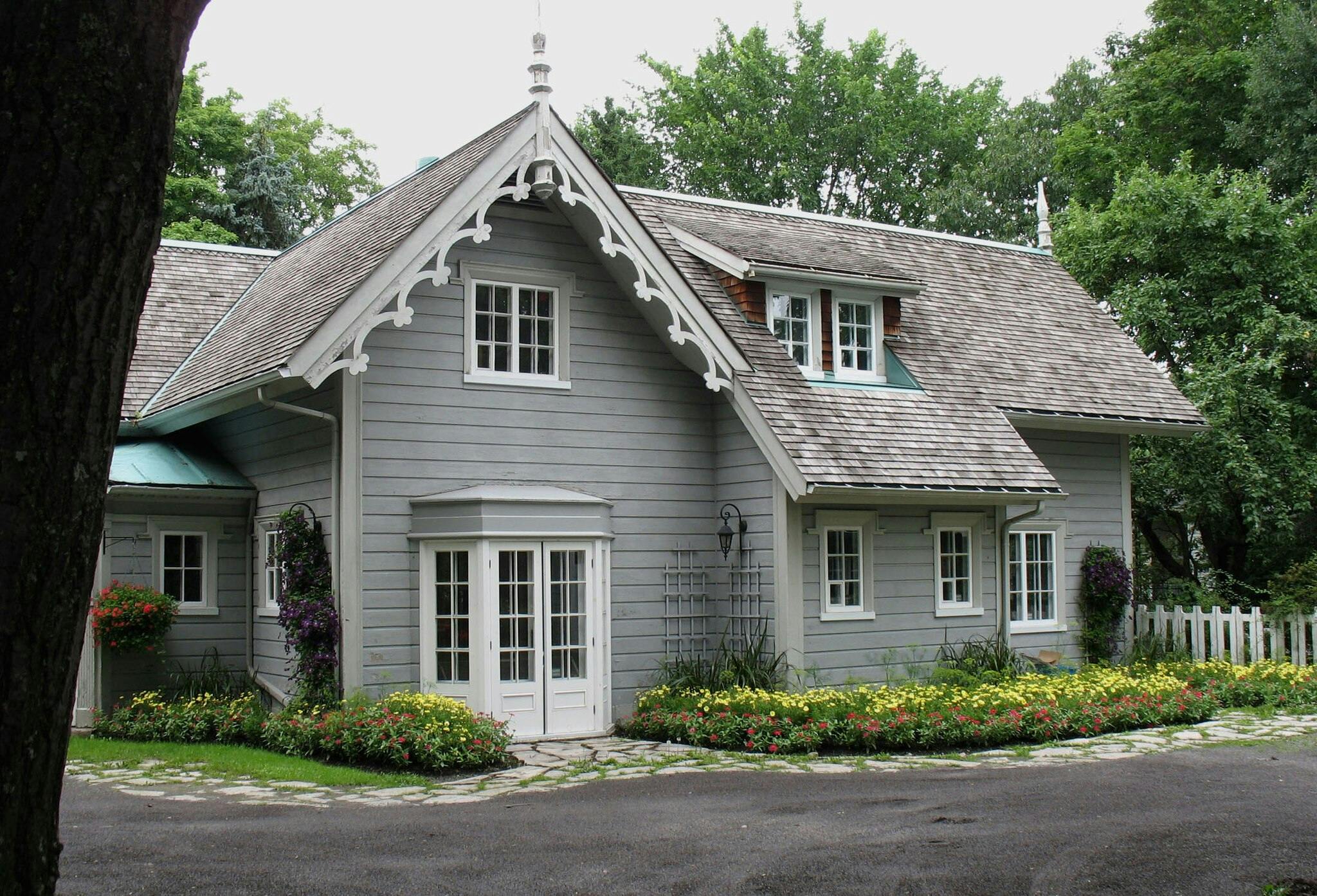 Ένα γοητευτικό γκρι εξοχικό σπίτι με λευκά διακοσμητικά στοιχεία, παρτέρια και έναν λευκό φράχτη, περιτριγυρισμένο από καταπράσινα δέντρα.