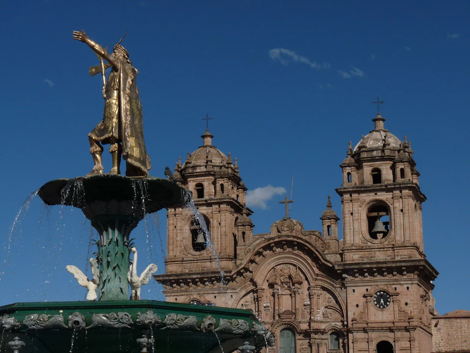 Bronze statue atop a water fountain in front of a historic cathedral with twin bell towers under a clear blue sky.