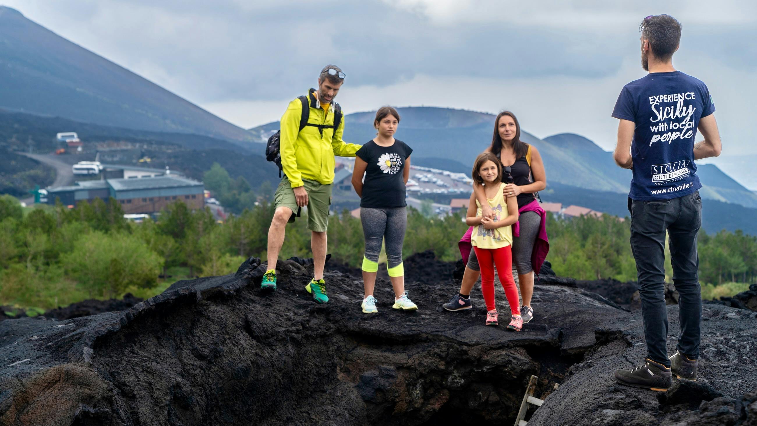 Een gezin van vijf staat op een rotsachtig terrein met in de verte heuvels en huizen zichtbaar onder een bewolkte hemel.