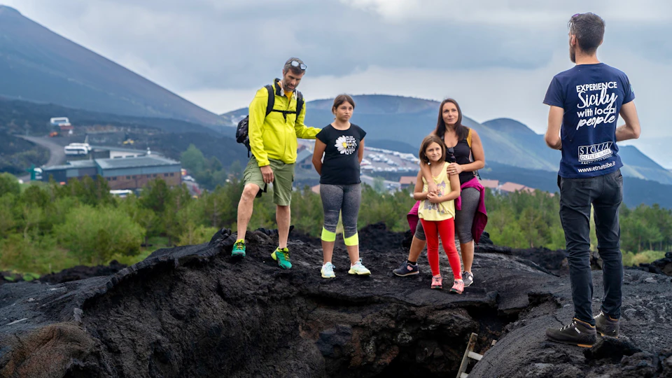 A family of five stands on a rocky terrain with distant hills and houses visible under a cloudy sky.