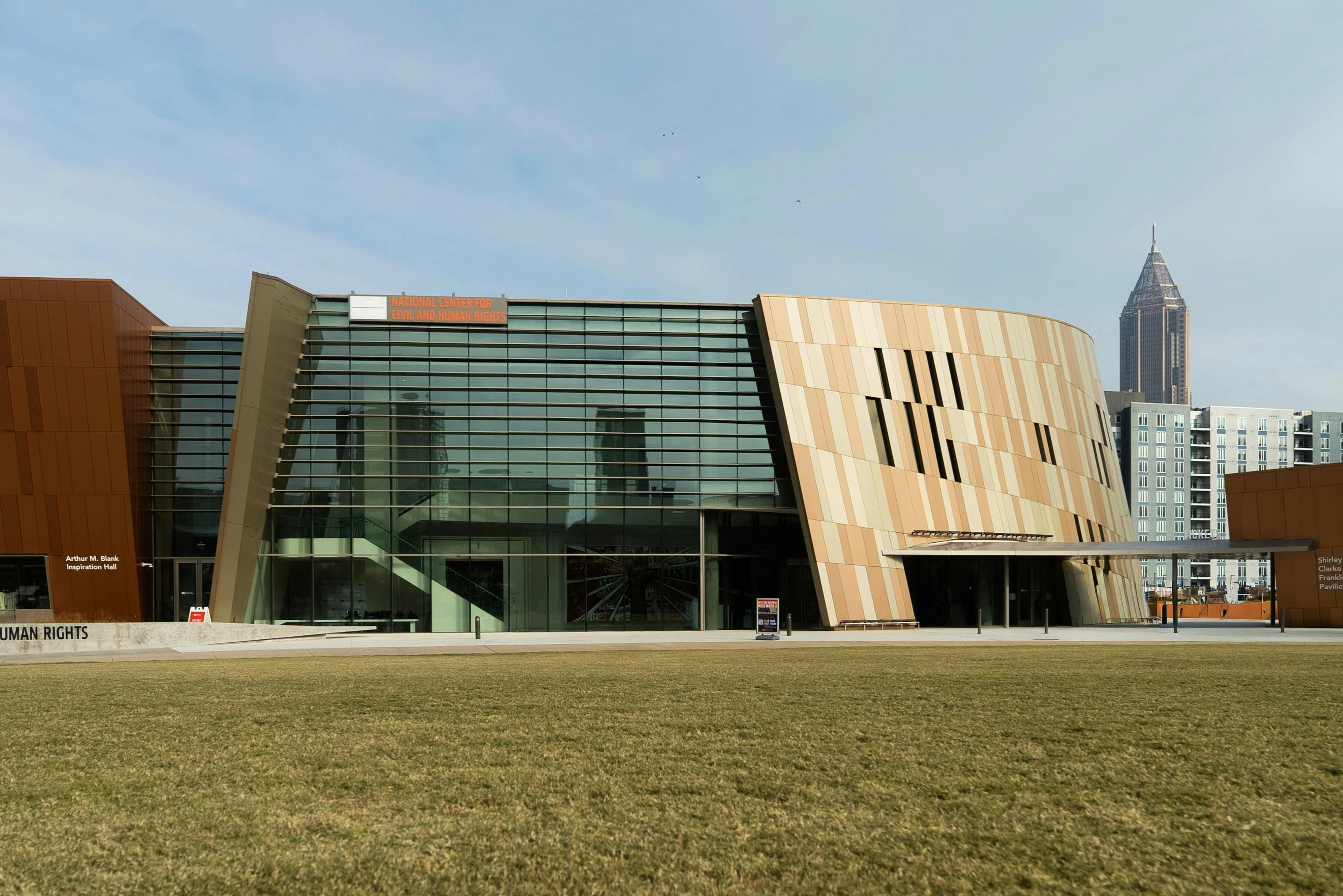 Edifício moderno com fachada de vidro e painéis de madeira, com um primeiro plano relvado e um céu limpo.