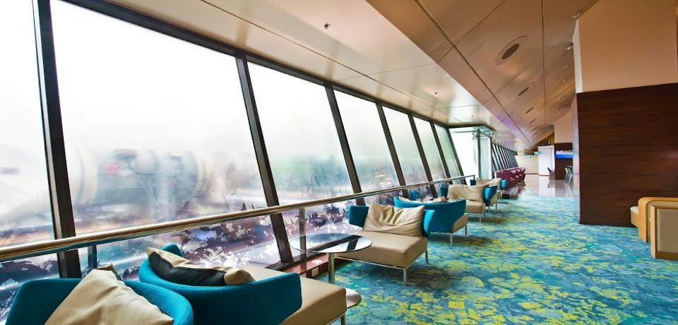 A modern lounge area with angled windows, blue armchairs, and small tables, featuring colorful patterned flooring.