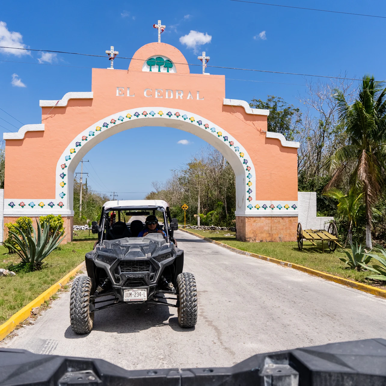 A black off-road vehicle passes under an arched entrance sign reading "El Cedral" on a sunny day. Small plants and trees line the path.