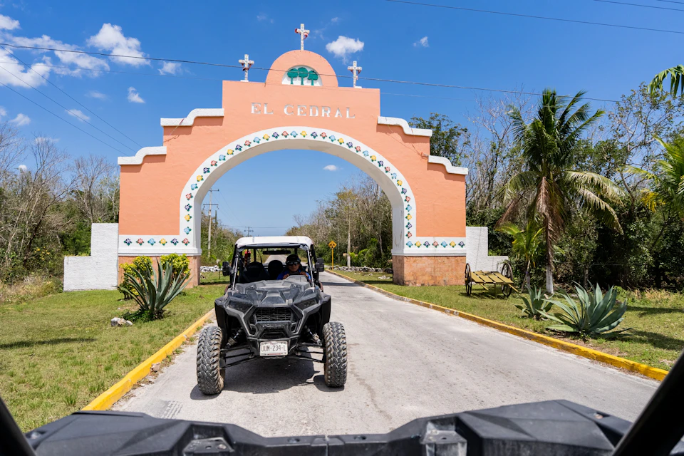 A black off-road vehicle passes under an arched entrance sign reading "El Cedral" on a sunny day. Small plants and trees line the path.