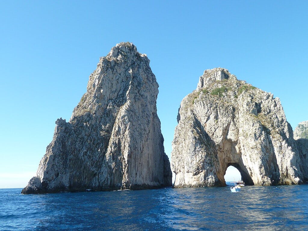 Two large rock formations rise from the ocean, one with an archway, under a clear blue sky. A small boat is visible in the water.
