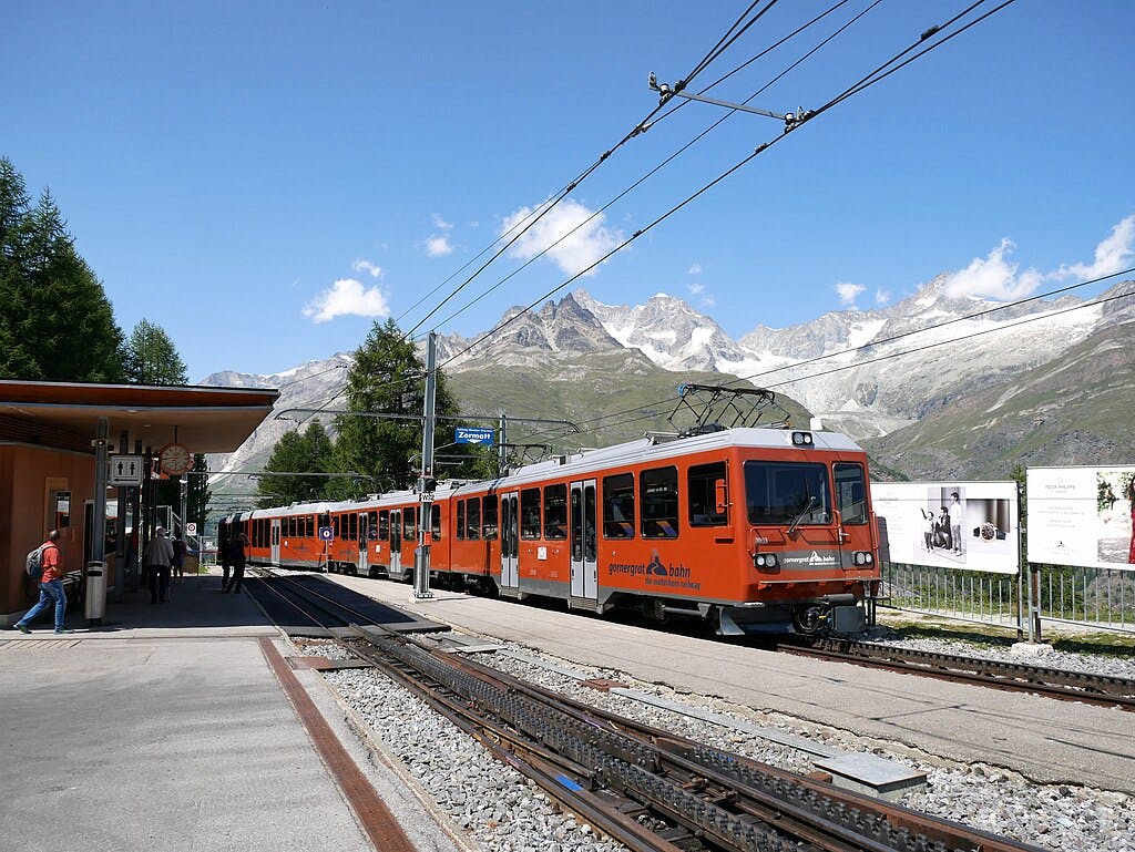 A red mountain train at the Gornergrat station with mountain peaks in the background and people waiting on the platform.