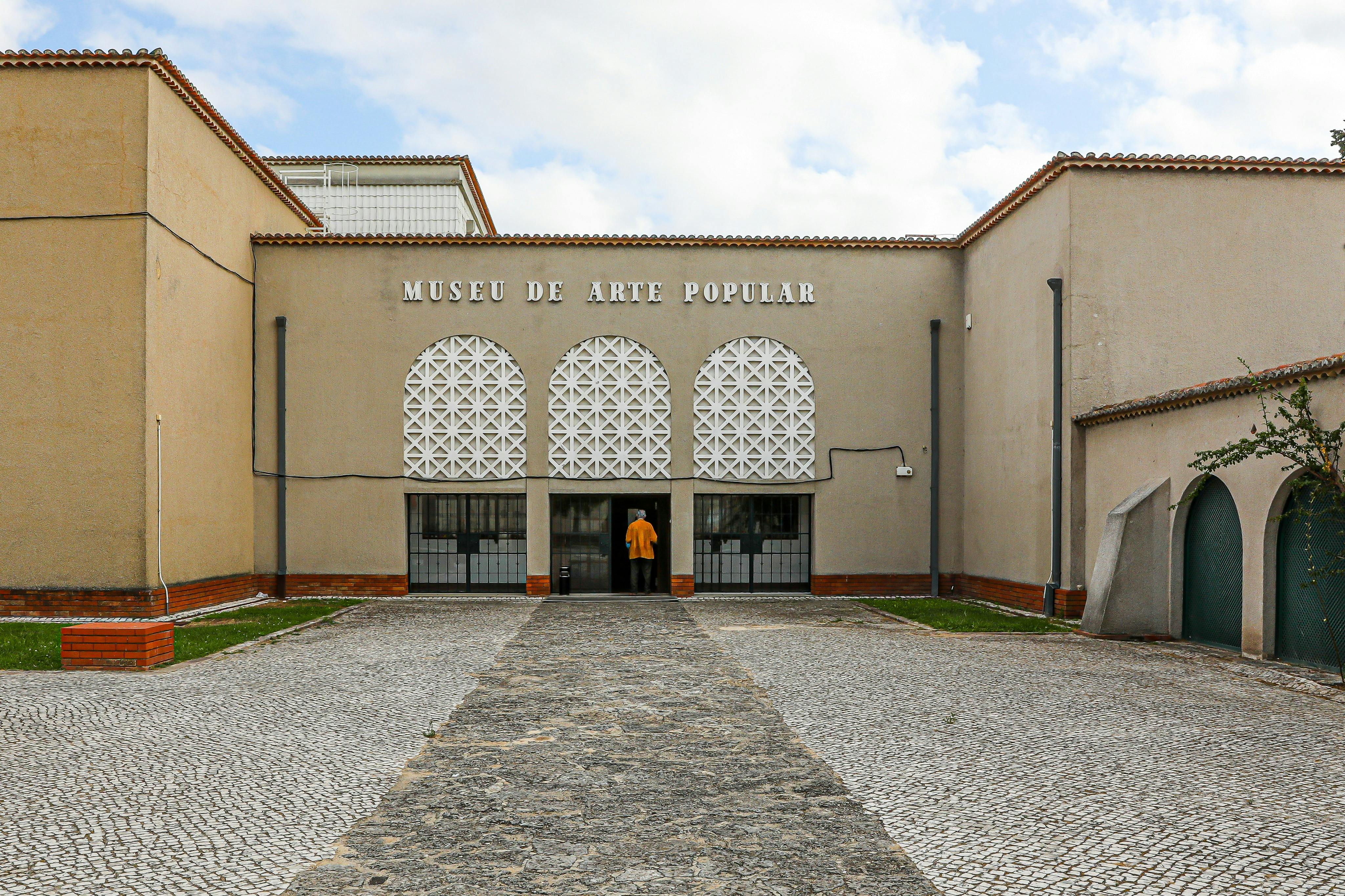 Une personne vêtue d'une chemise jaune entre dans le Museu de Arte Popular, un bâtiment beige avec des fenêtres à motifs et un chemin pavé de pierres.