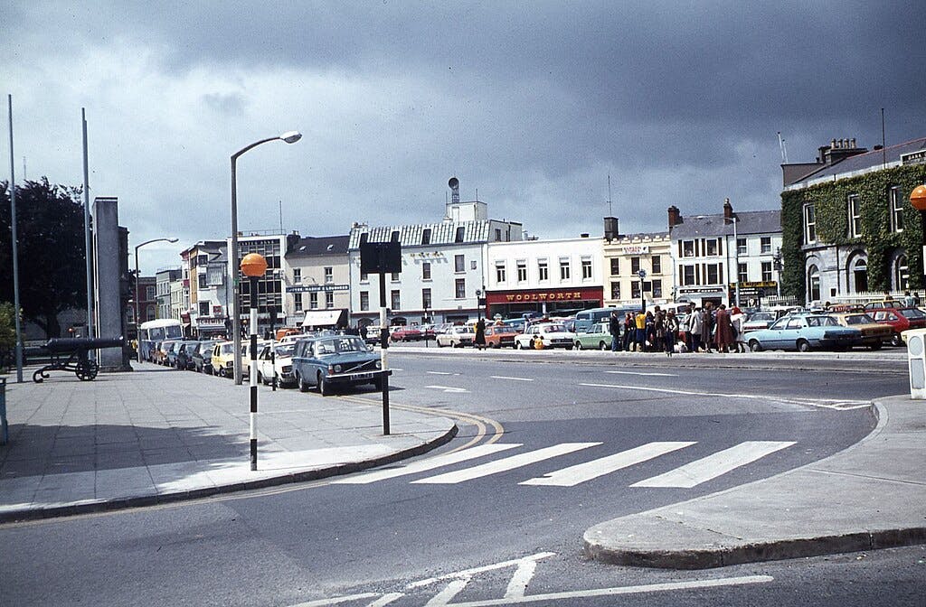 Уличная сцена с припаркованными автомобилями, группой людей на тротуаре и зданиями, включая магазин Woolworth под пасмурным небом.