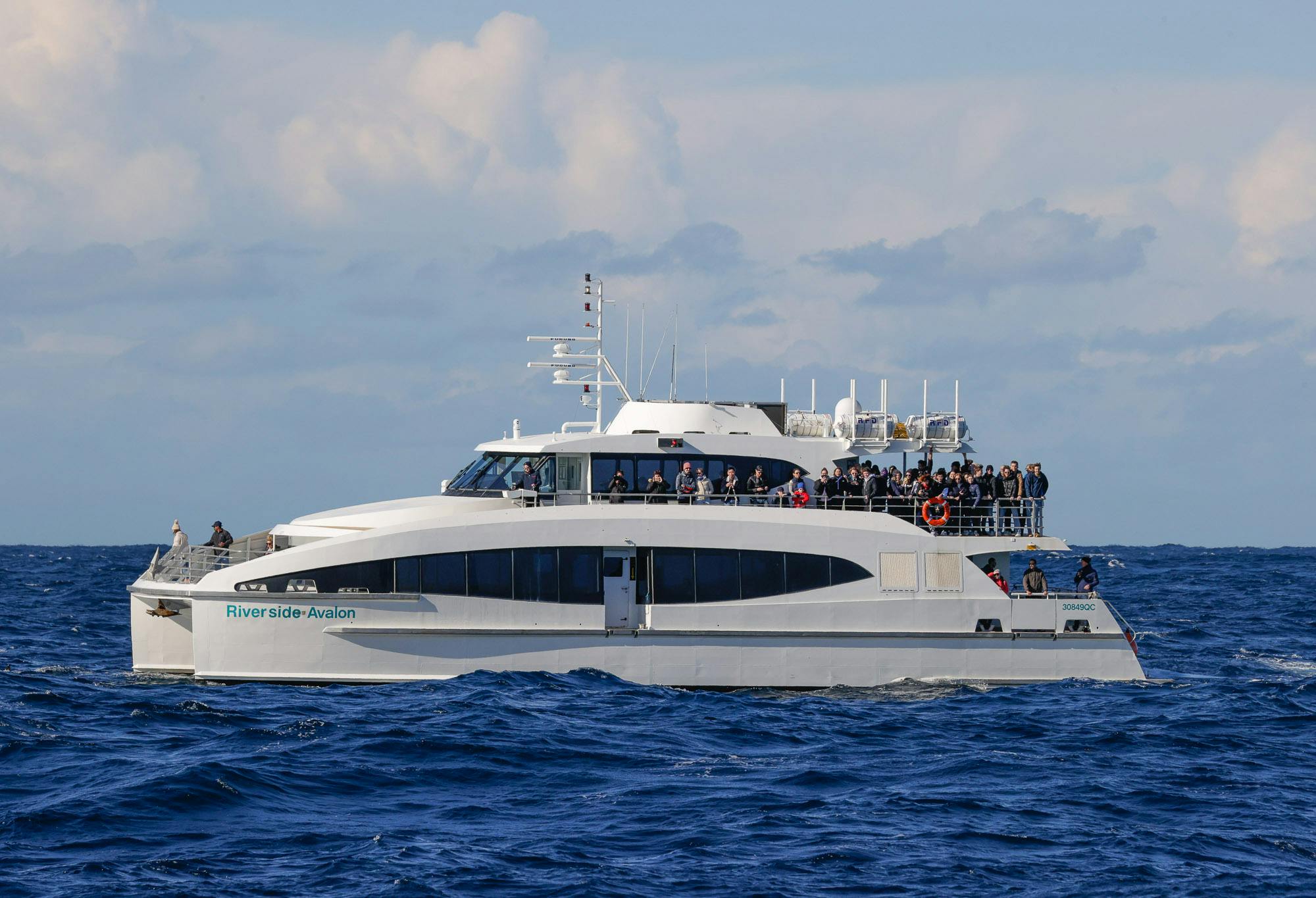 A passenger ferry with people onboard sails on the ocean under a cloudy sky.