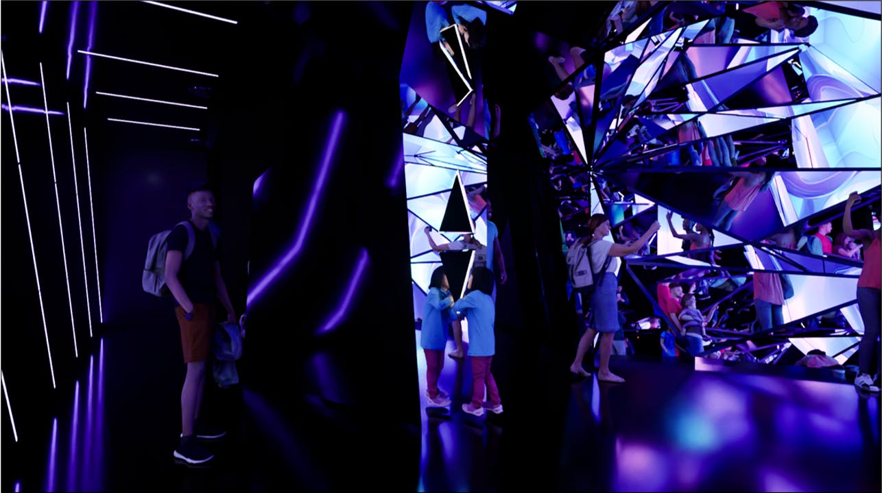 La gente recorre una exposición inmersiva llena de espejos, con iluminación morada y reflejos geométricos.