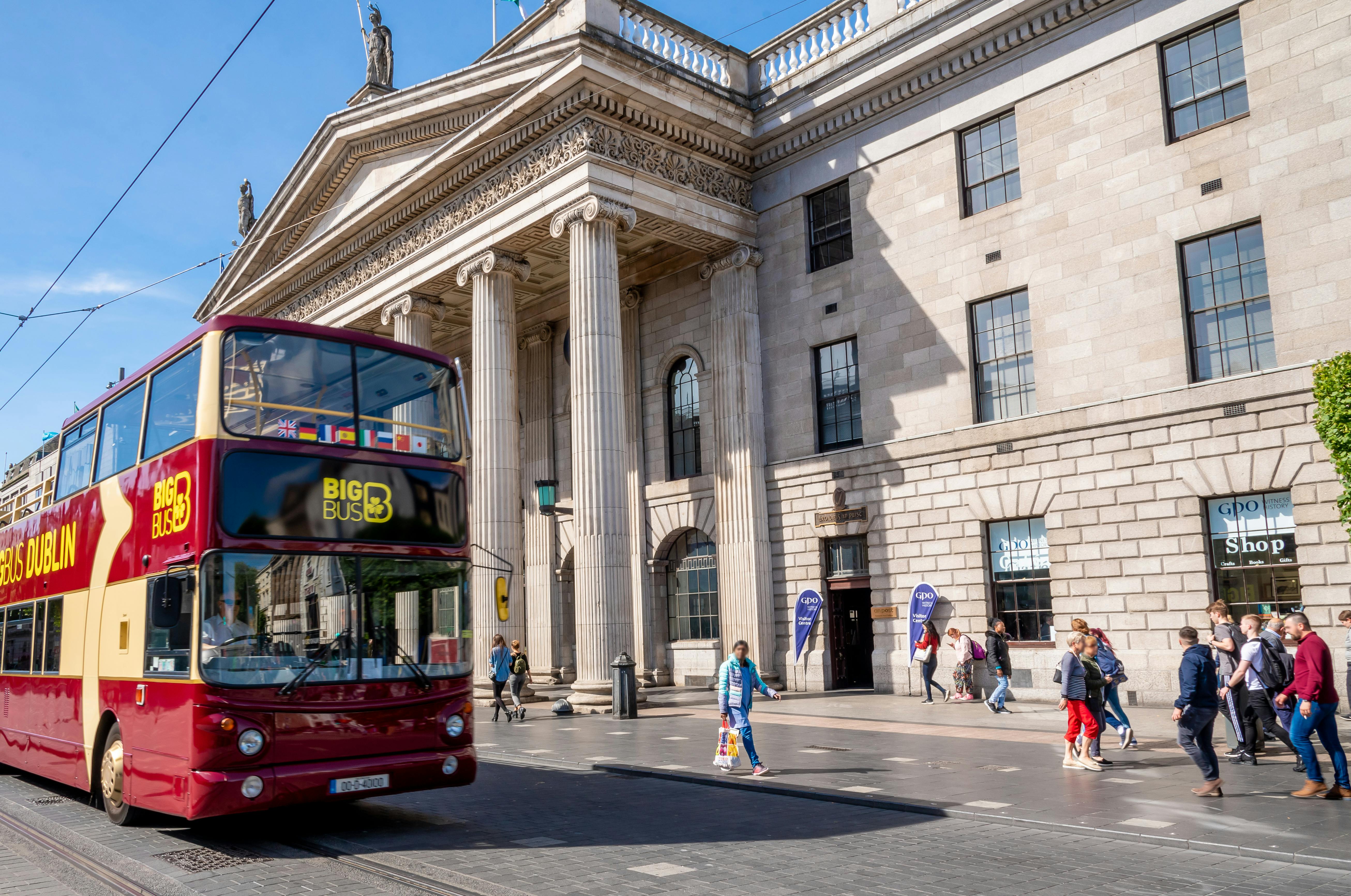 Ένα κόκκινο και κίτρινο διώροφο τουριστικό λεωφορείο περνάει από ένα ιστορικό κτίριο με κίονες. Οι πεζοί περπατούν στο πεζοδρόμιο.