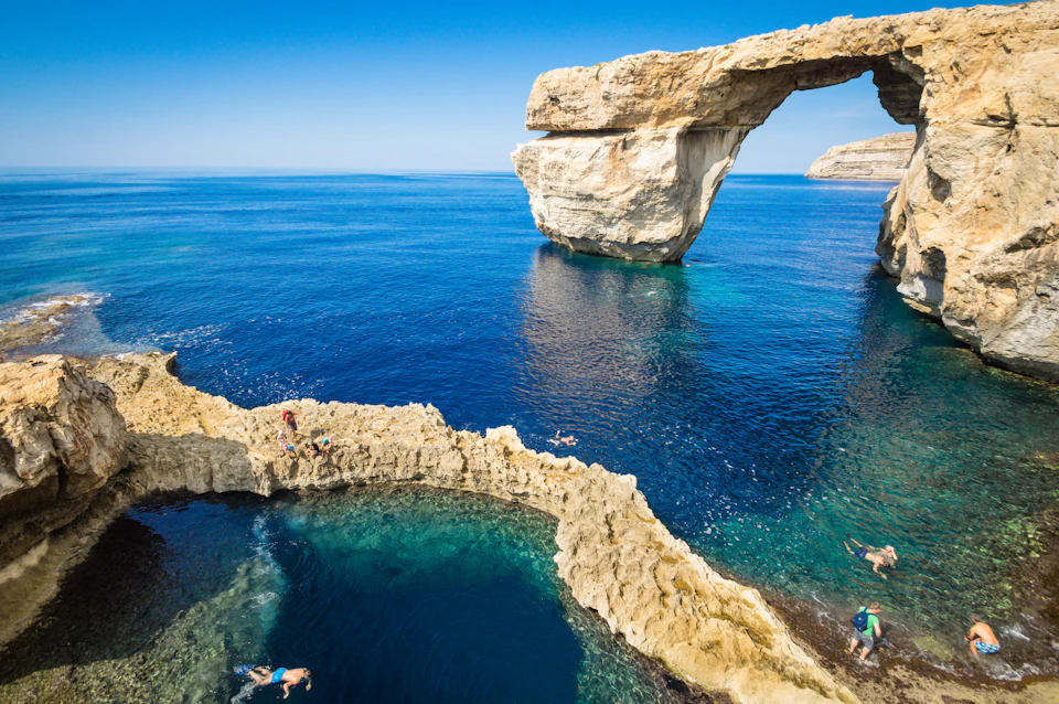 People swimming and sunbathing on rocky formations by a natural arch over clear blue sea under a clear sky.