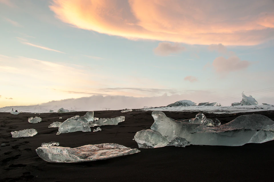 Chunks of clear ice scattered on a black sand beach under a sky with soft orange and pink clouds.