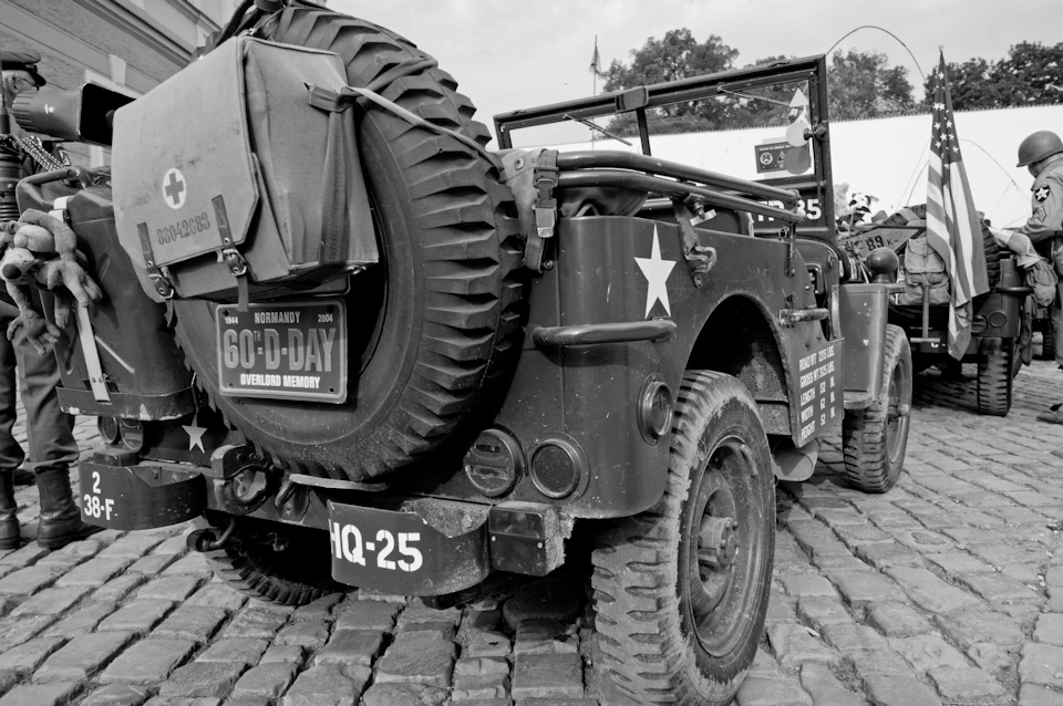 A vintage military jeep with a rear-mounted spare tire and D-Day commemorative signs on a cobblestone street.