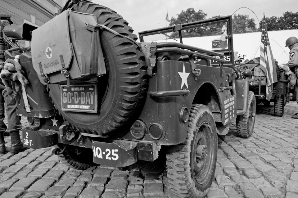 A vintage military jeep with a rear-mounted spare tire and D-Day commemorative signs on a cobblestone street.