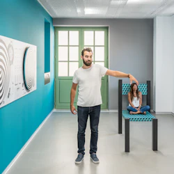 En man står och pekar medan en sittande kvinna i bakgrunden ser liten ut och skapar en optisk illusion i ett modernt rum med konstverk.