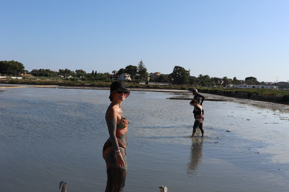 A woman in swimwear and a hat stands in shallow water, while two others wade in the background near a rural landscape.