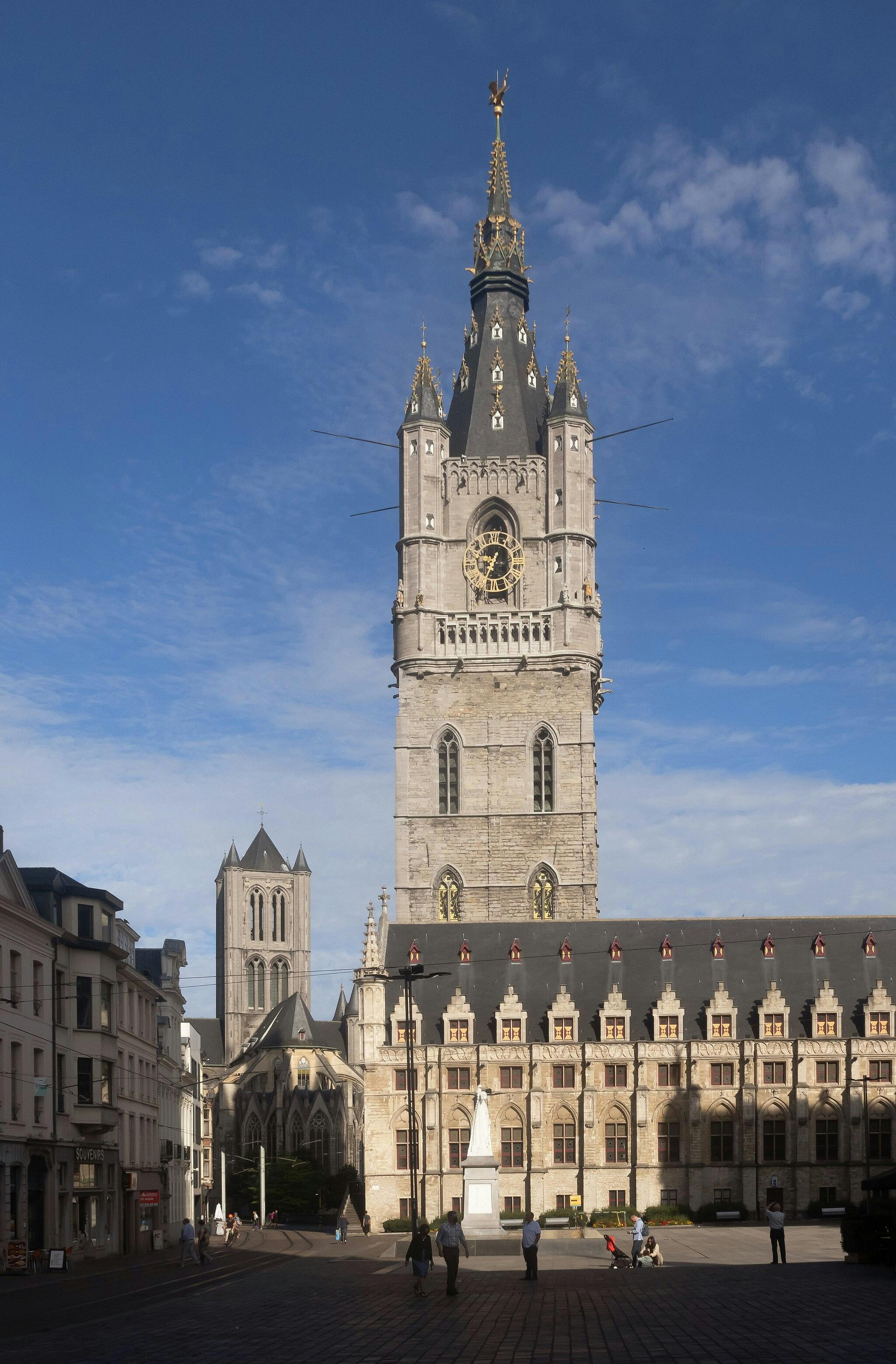 Grande tour d'horloge de style gothique avec une girouette dorée, bâtiments historiques adjacents et personnes marchant sur une place éclairée par le soleil.