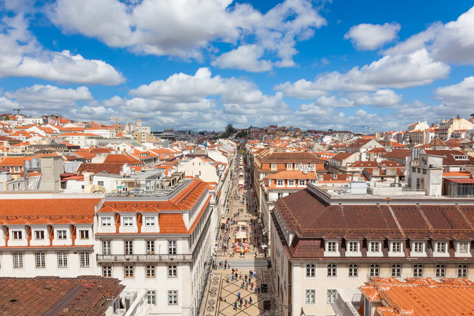 Aerial view of a bustling pedestrian street lined with buildings featuring red-tiled roofs under a partly cloudy blue sky.