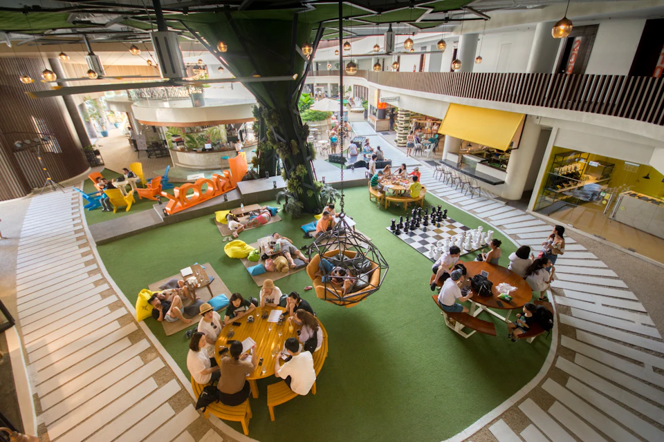 Open indoor space with green flooring, tables, a large tree-like structure, people sitting, and a chessboard on the side.