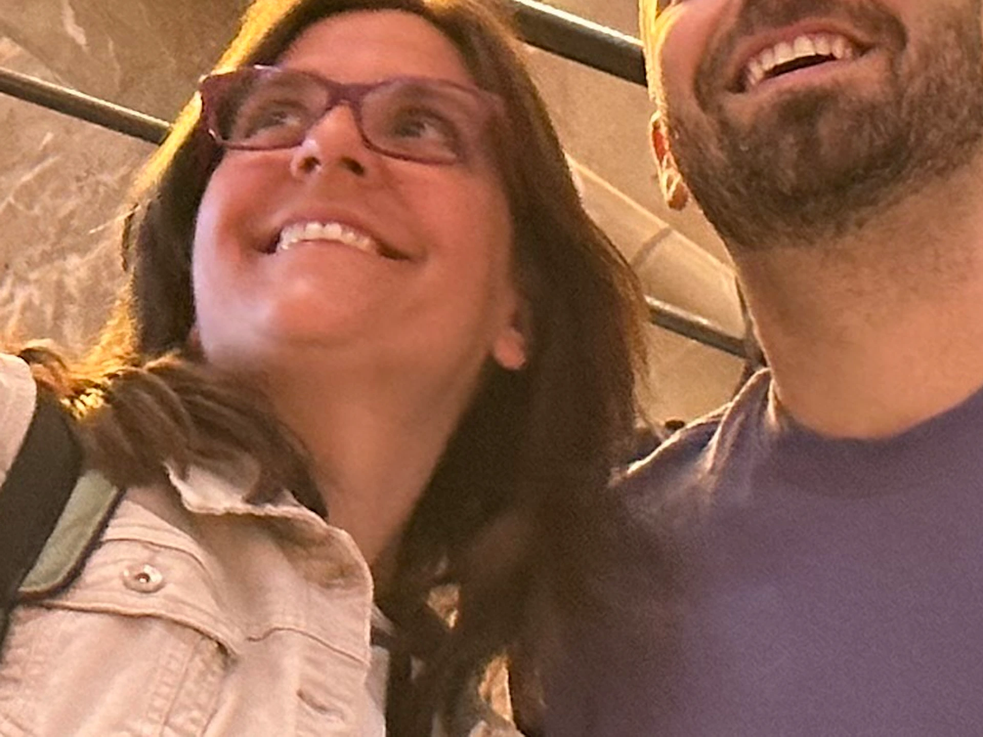 Two people standing and smiling inside a building with ornate architectural details and a marble staircase. One points upward.