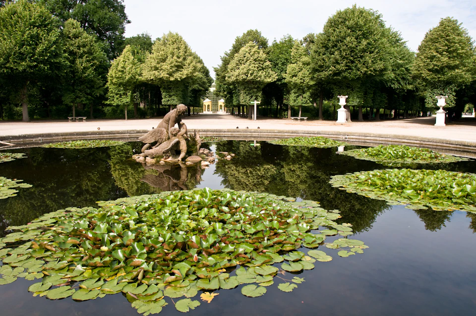 A fountain with lily pads and a statue in the center, surrounded by dense trees and a distant arched entrance.