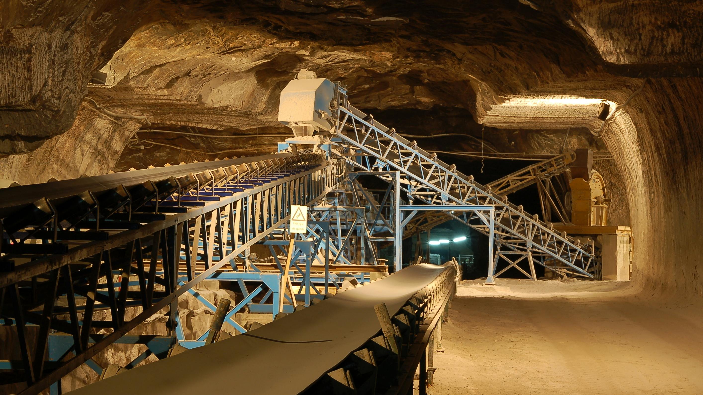 Underground mining operation with conveyor belts, machinery, and rock walls. Dim lighting with overhead lights.