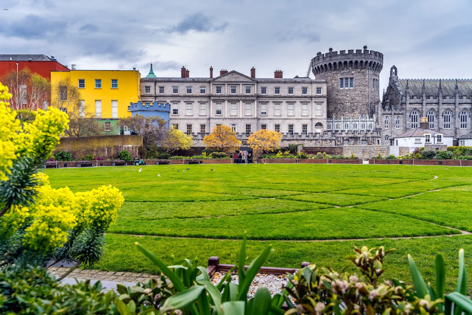 A grassy courtyard with trimmed hedges and flowerbeds, bordered by a historic building and tower under a cloudy sky.