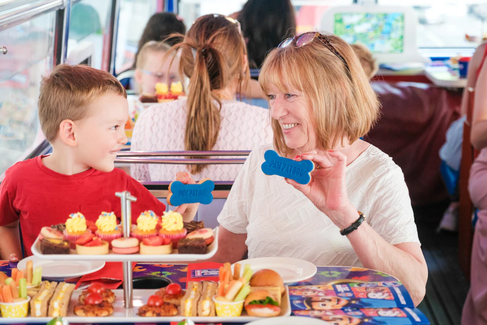 A woman and a boy sit at a table with cupcakes, burgers, and sweets, smiling and holding blue biscuits shaped like bones.