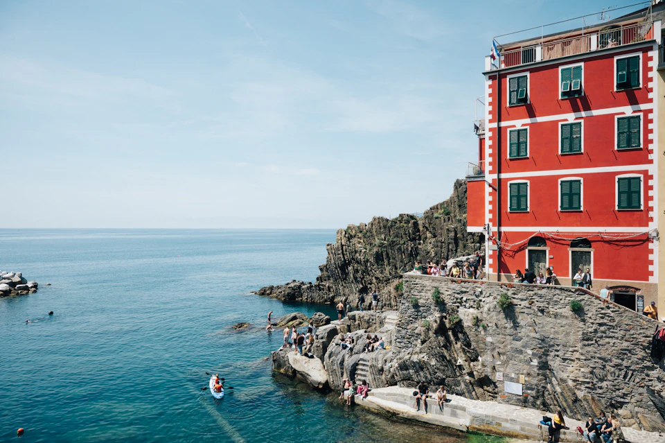 People enjoying the water near rocky cliffs and a red building by the sea on a clear day. A boat with a person is nearby.