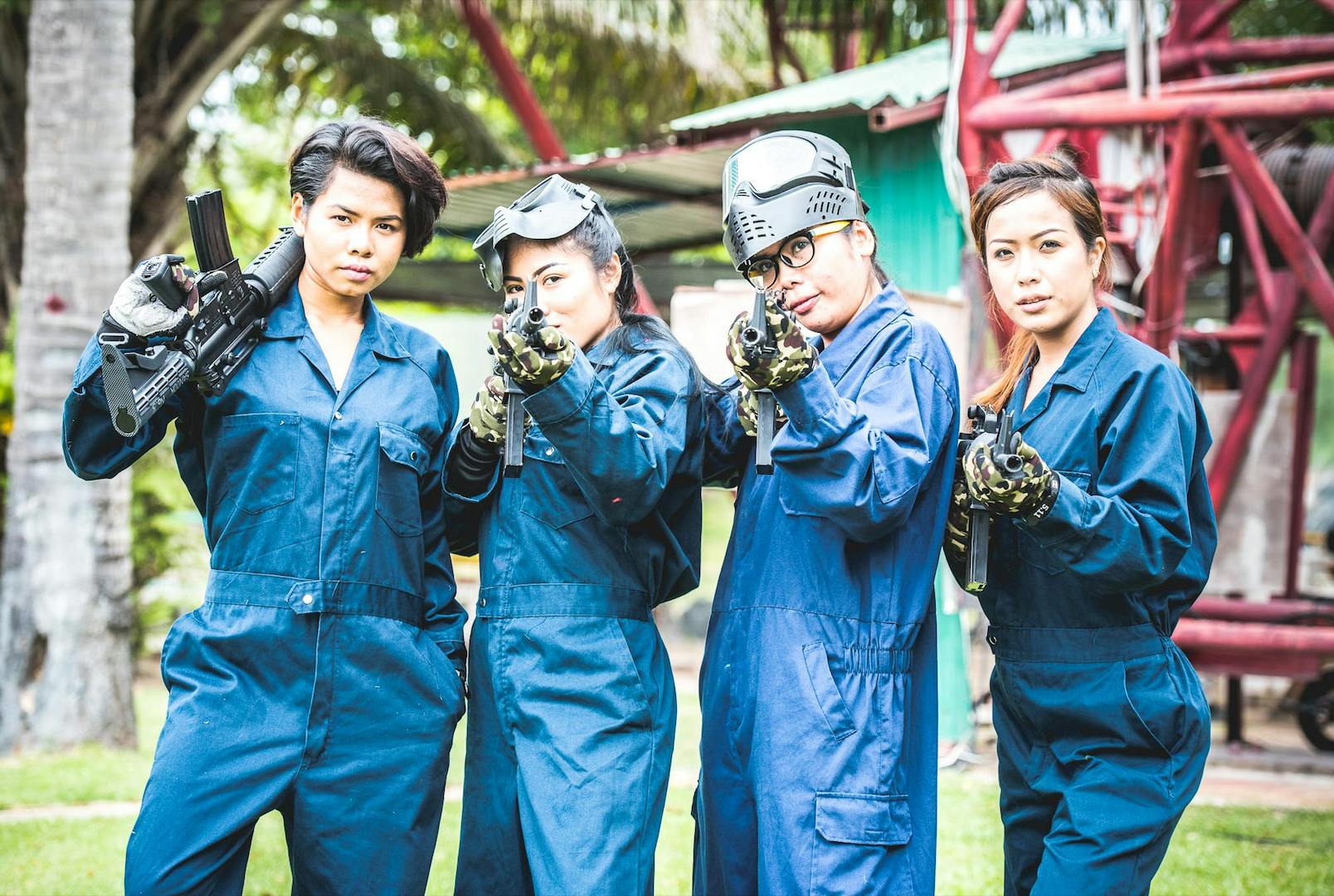 Cztery osoby w niebieskich kombinezonach trzymające pistolety do paintballa, stojące na zewnątrz z zielono-czerwoną strukturą w tle.