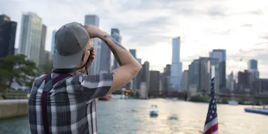 Una persona su una barca fotografa un paesaggio urbano con alti edifici; una bandiera americana si trova sulla poppa della barca.
